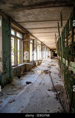 Ukraine, Pripyat: Langer, verfallener Flur innerhalb einer verlassenen Schule mit abblätternder Decke und Wänden, grünen Metallgeländern, offenen Fenstern mit Vegetati Stockfoto