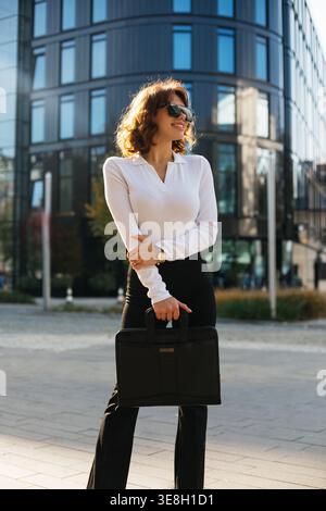 Frau steht draußen mit Handtasche. Attraktive Geschäftsfrau in elegantem Outfit und Sonnenbrille. Professioneller Firmenstil. Urbanes Lifestyle und Bus Stockfoto
