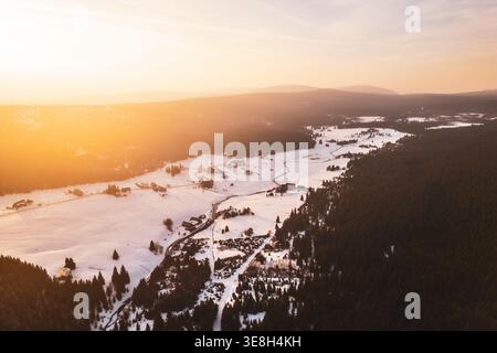 Die Szene zeigt das Bergdorf Jizerka, das im Winter mit Schnee bedeckt ist. Eine Drohne fängt das Dorfbild, die Bäume und das schneebedeckte Land unter einem Sonnenuntergang ein. Stockfoto
