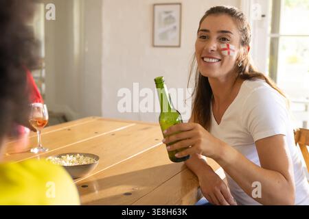 Grüne Bierflasche sitzt auf hölzernem Esstisch neben dem Fenster mit Popcorn-Schüssel und Weinglas Stockfoto