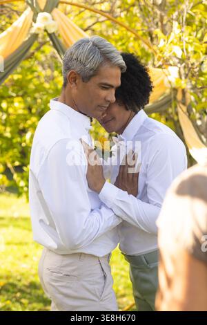 Männliche Partner umarmen sich unter einem Holzbogen im Garten, tragen weiße Hemden und Boutonniere Stockfoto