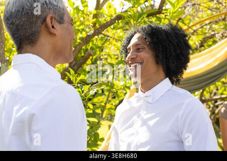 Zwei Männer, die bei der Zeremonie feiern, beginnen im Garten mit drapiertem Stoff, weißen Hemden, Sonnenlicht auf Gesichtern Stockfoto