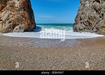 Flacher Blick auf türkisfarbene Meereswellen, die zwischen massiven Kalksteinfelsen auf einen Kiesstrand unter klarem blauen Himmel mit viel Kopierraum krachen Stockfoto
