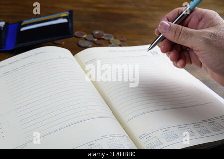 Hand halten Sie den Stift über der leeren Planerseite mit Brieftasche und Münzen im Hintergrund, bereit für die Budgetierung oder Notizen. Stockfoto