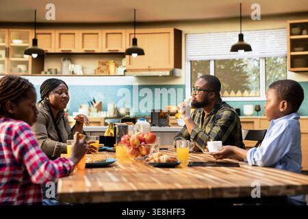 Afroamerikanische Familie teilt Gebäck und Getränke zum Frühstück zu Hause, Eltern servieren Rührei, Toast und Obst. Gemütlicher Moment in der Küche fängt Wärme, Entspannung und fröhliche Zweisamkeit ein. Stockfoto