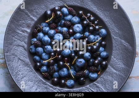 Draufsicht des Beerensortiments in schwarzer Platte auf strukturiertem Hintergrund. Heidelbeeren und Maulbeeren, minimale Zusammensetzung Stockfoto