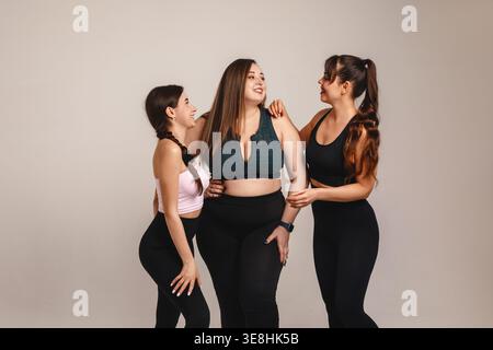 Fröhliches Lifestyle-Konzept. Glückliche Frauen, die mit einem kurvigen Model in großer Größe befreundet sind, lachen gemeinsam in Studiosportbekleidung. Verschiedene weibliche Gruppen teilen Freundschaft, Stockfoto