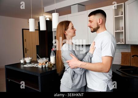 Ein Mann und eine Frau stehen dicht in einer Küche und schauen sich gegenseitig in die Augen. Sie sind in einem warmen Moment beschäftigt, während sie zusammen Speisen zubereiten. Die Küche Stockfoto
