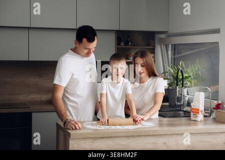 Eine Familie genießt eine gute Zeit in der Küche. Ein Kind rollt mit Hilfe beider Eltern Teig auf einem Tisch. Sie haben Spaß zusammen, während sie lächeln Stockfoto