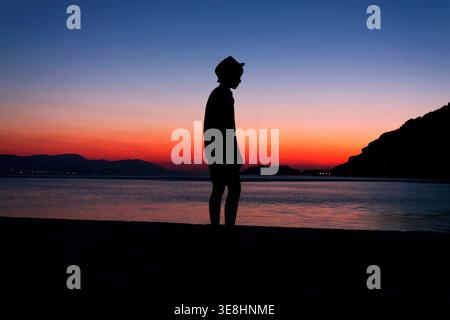 Silhouette einer Person mit Hut am Strand bei farbenfrohen Sonnenuntergang in Griechenland Stockfoto