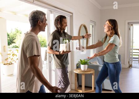 Drei Freunde grüßen in hellem Wohnzimmer in der Nähe der Schiebetür, Mann hält Pappe Bierträger Stockfoto