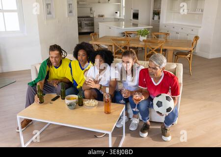 Verschiedene Freunde, die sich nach vorne lehnen und Fußballspiele im hellen Wohnzimmer mit Fußball beobachten Stockfoto