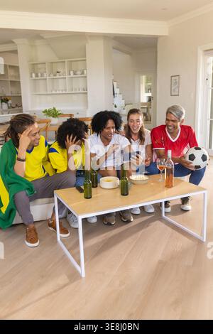 Verschiedene Freunde, die Fußballtrikots tragen, schauen sich das Spiel zu Hause mit einem Fußballbier an Stockfoto