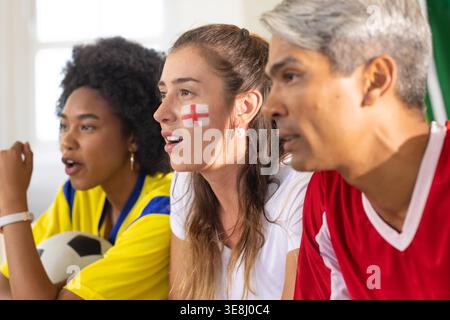 Verschiedene Fans, die Fußballspiele auf der Couch, in Trikots, in Ballhaltung und rotem Kreuz beobachten Stockfoto