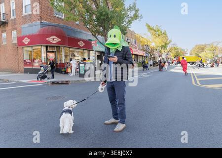 Ein Mann in einer grünen Entenkostüm-Maske überprüft sein Handy, während er mit seinem weißen Hund unterwegs ist während eines Halloween-Straßenfestes in Leonia, NJ Stockfoto