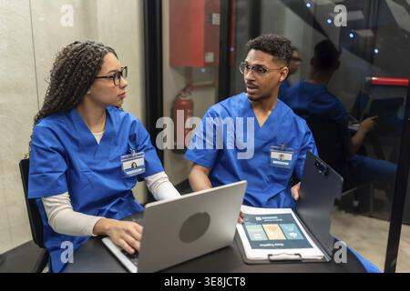Medizinisches Personal in blauem Scrubs diskutiert Patienteninformationen, arbeitet an einem Laptop zusammen und überprüft Dokumente in einem modernen Krankenhaus oder einer Klinik Stockfoto
