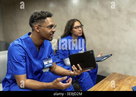 Medizinische Fachkräfte in Blue Scrubs arbeiten zusammen und diskutieren Patientendaten, nutzen digitale Tablets für effiziente Kommunikation und moderne ME Stockfoto
