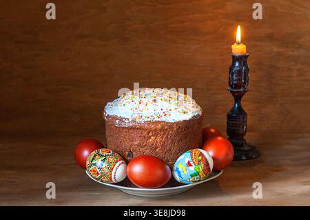 Traditionelles Osterstilleben mit Kulich-Kuchen mit Glasur und Streuseln, bunt bemalten Eiern, beleuchteten Kerzen und blühenden Zweigen in weißer Grube Stockfoto