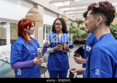 Gruppe von verschiedenen Studenten im Gesundheitswesen in blauen Peelings, die zusammen stehen, lächeln und unterhalten, während sie Kaffeetassen und eine Wasserflasche während einer Re halten Stockfoto