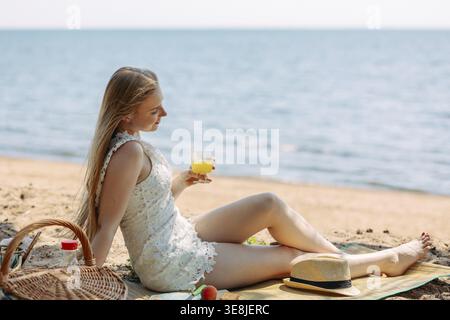 Romantische blonde Frau mit Strohhut beim Picknick am Strand. Frisches Obst, Saft, Kekse und Pfirsiche. Stockfoto