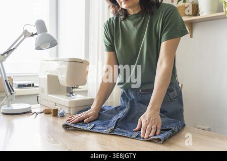 Frau, die an einem Schreibtisch steht, eine Jeans glättet, sich mit einer Nähmaschine in der Nähe auf das Nähen vorbereitet oder verändert. Dinge wiederverwenden. Umwelt Stockfoto
