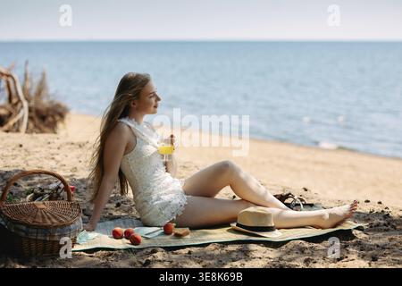 Romantische blonde Frau mit Strohhut beim Picknick am Strand. Frisches Obst, Saft, Kekse und Pfirsiche. Stockfoto