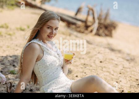 Romantische blonde Frau mit Strohhut beim Picknick am Strand. Frisches Obst, Saft, Kekse und Pfirsiche. Stockfoto