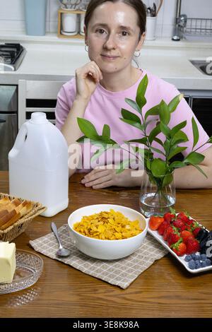 Junge Frau, die Milch in eine Schüssel Cornflakes mit frischen Beeren auf einem Holztisch in einer gemütlichen Küche gießt. Gesundes Frühstück Stockfoto