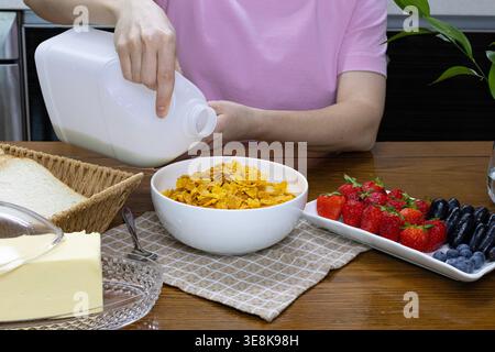 Junge Frau, die Milch in eine Schüssel Cornflakes mit frischen Beeren auf einem Holztisch in einer gemütlichen Küche gießt. Gesundes Frühstück Stockfoto