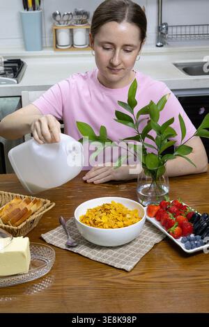 Junge Frau, die Milch in eine Schüssel Cornflakes mit frischen Beeren auf einem Holztisch in einer gemütlichen Küche gießt. Gesundes Frühstück Stockfoto