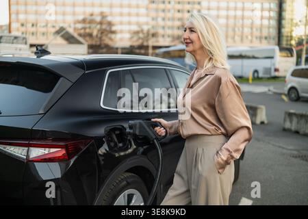 Eine blonde Frau greift mit einer Ladestation eines Elektrofahrzeugs in Berührung, während sie den Prozess genießt. Stockfoto