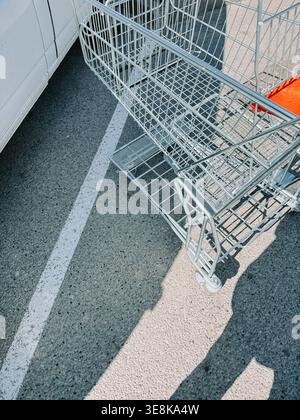 Verlassener Einkaufswagen aus Metall, der auf einem markierten Parkplatz neben einem Fahrzeug steht, wirft einen starken Schatten. Visuelles Konzept von unvorsichtigem Verhalten, Par Stockfoto
