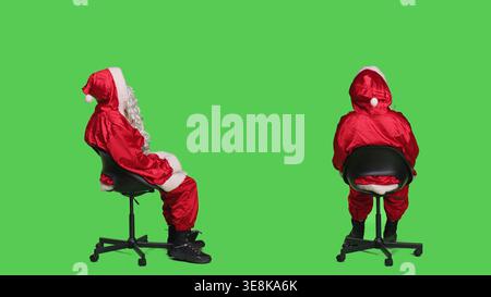 Vater weihnachten Cosplay sitzt auf einem Stuhl, schaut sich im Studio um und trägt festliches rotes Kostüm mit Hut und Bart. Die berühmte dezemberfigur posiert auf dem Ganzkörper-Greenscreen-Hintergrund. Stockfoto