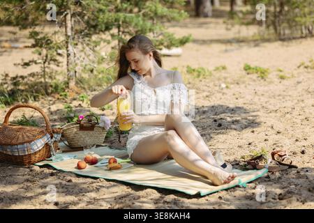 Romantische blonde Frau mit Strohhut beim Picknick am Strand. Frisches Obst, Saft, Kekse und Pfirsiche. Stockfoto