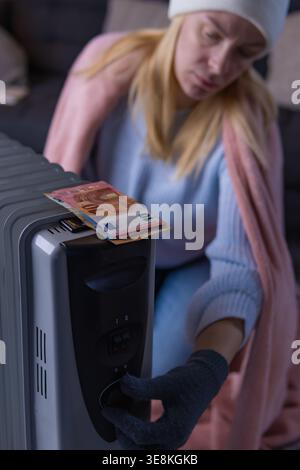Frau mit warmer Kleidung und Handschuhen, die eine elektrische Heizung einstellen, wobei die steigenden Stromrechnungen, Energiearmut und die finanzielle Belastung durch Wärme hervorgehoben werden Stockfoto