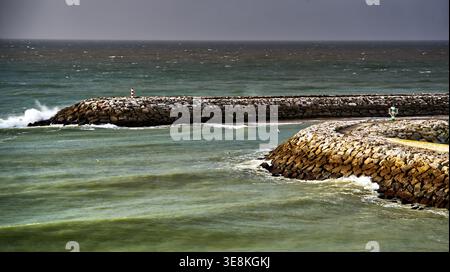 Ozeanwellen stürzen gegen die felsigen Wellenbrecher des Jachthafens von albufeira, mit einem kleinen Leuchtturm, der den Eingang unter einem glühenden Himmel in portugal markiert. Stockfoto