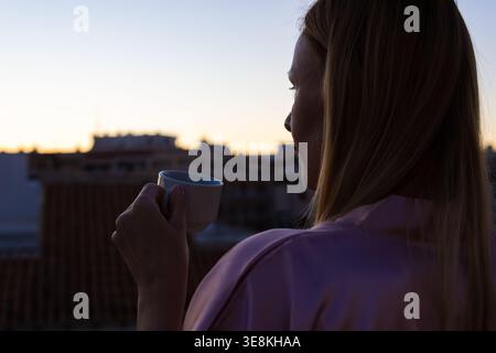 Frau, die auf einem Balkon steht, eine Kaffeetasse hält, auf die Skyline der Stadt blickt, während die Sonne aufgeht, und einen Moment der persönlichen Reflexion und Entspannung genießt Stockfoto