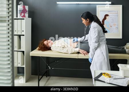 Weitaufnahme einer Ärztin, die schwangere kaukasische Frau mit Stethoskop während der pränatalen Untersuchung in der Klinik untersucht. Unterstützung der Geburtshilfe, geburtshilfliches Marketing Stockfoto