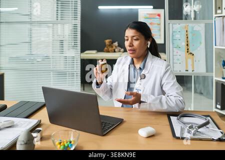 Hispanische junge Ärztin mit Arzneiflasche und Sprechpatientin auf Laptop während Telehealth-Termin. Unterstützung der Online-Gesundheitsversorgung, Ferndiagnose Stockfoto