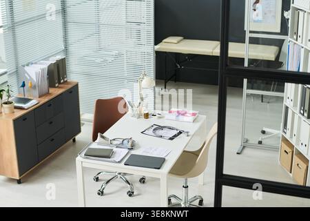Weitwinkelaufnahme einer leeren medizinischen Praxis, die einen Schreibtisch mit Stethoskop, Tablet, Dokumente, Untersuchungsbereich zeigt, was einen Arbeitsablauf für die medizinische Beratung vorschlägt Stockfoto