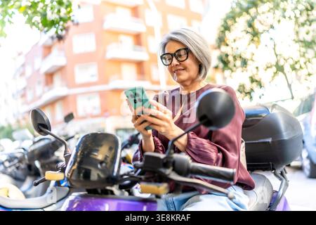 Ältere Frau, die in der Stadt ihr Smartphone auf dem Roller benutzt Stockfoto