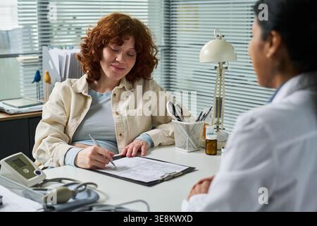 Junge Frau, die ein medizinisches Formular während der Konsultation mit einer Ärztin unterschreibt, Behandlung oder Kontrollbedürfnisse bespricht. Nützlich für medizinische Zustimmung, Patientenaufnahme, Versicherung, klinisches Marketing Stockfoto