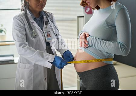 Mittlere Aufnahme von schwangeren jungen erwachsenen Frauen, die Bauchmessung von weiblichen Ärzten während der pränatalen Untersuchung erhalten, Unterstützung der Mutterschaft Pflege, geburtshilfliches Gesundheitsmarketing Stockfoto