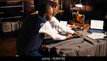 afroamerikanische Schneider schneiden Kleidungsstücke und arbeiten im Team an maßgeschneiderten Kleidungsstücken. Handwerker und Assistent mit Schneidewerkzeugen im Atelier, Vorbereitung der Handarbeit. Handgeführte Aufnahme. Stockfoto