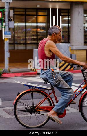 Taipei, Taiwan - 22. April 2024: Ein älterer Mann in rotem Tanktop und Jeans fährt auf einer belebten Stadtstraße Taipeis mit dem Fahrrad, inmitten von Straßenverkehr und Stadtgebäuden. Stockfoto