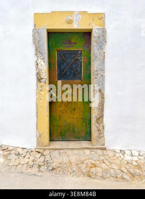 Verwitterte grüne Metalltür mit einem kleinen eisernen Fenster in einem gelben und Steinrahmen auf einer weißen Wand über einem Kopfsteinpflasterweg algarve portugal Stockfoto