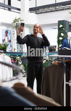 Der arabische Influencer wirbt für Boutiquen, während er im Bekleidungsgeschäft mit dem Smartphone streamt. Shopping Mall junger Kunde hält Handy, winkt hoch und nimmt Video auf Stockfoto