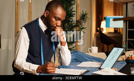 afroamerikanischer Schneider zeichnet Kleidung auf Papier und erstellt Skizzen, bevor er die Kleidungslinie handgefertigt entwirft. Couturier arbeitet an einer neuen Modekollektion im Atelier mit Werkzeugen. Stockfoto