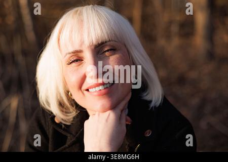 Ein detailgetreues Porträt einer schönen jungen Frau, die ihr Kinn auf der Hand hält und in den Strahlen der warmen Frühlingssonne lächelt. Lächelnde Blondine. Stockfoto