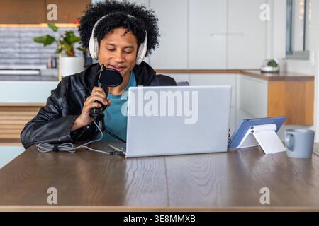 Afroamerikanischer Mann mit Headset, der in der Küche sitzt und ein Mikrofon mit Popfilter mit Laptop hält. Creator, Podcast, Streaming, Kopfhörer, Studio, Arbeitsbereich, Tablet Stockfoto